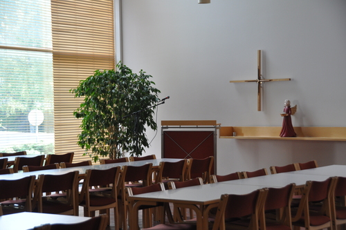 Liedon seurakuntatalon Mestarinsalin yleisnäkymässä pöytäryhmiä, puhujankoroke, osa seinäalttaria sekä risti seinällä. 