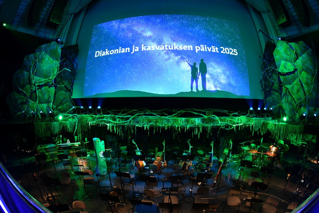 Hämärässä salissa diakonian ja kasvatuksen päivien dia seinällä ja edustalla orkesterin soittimia.