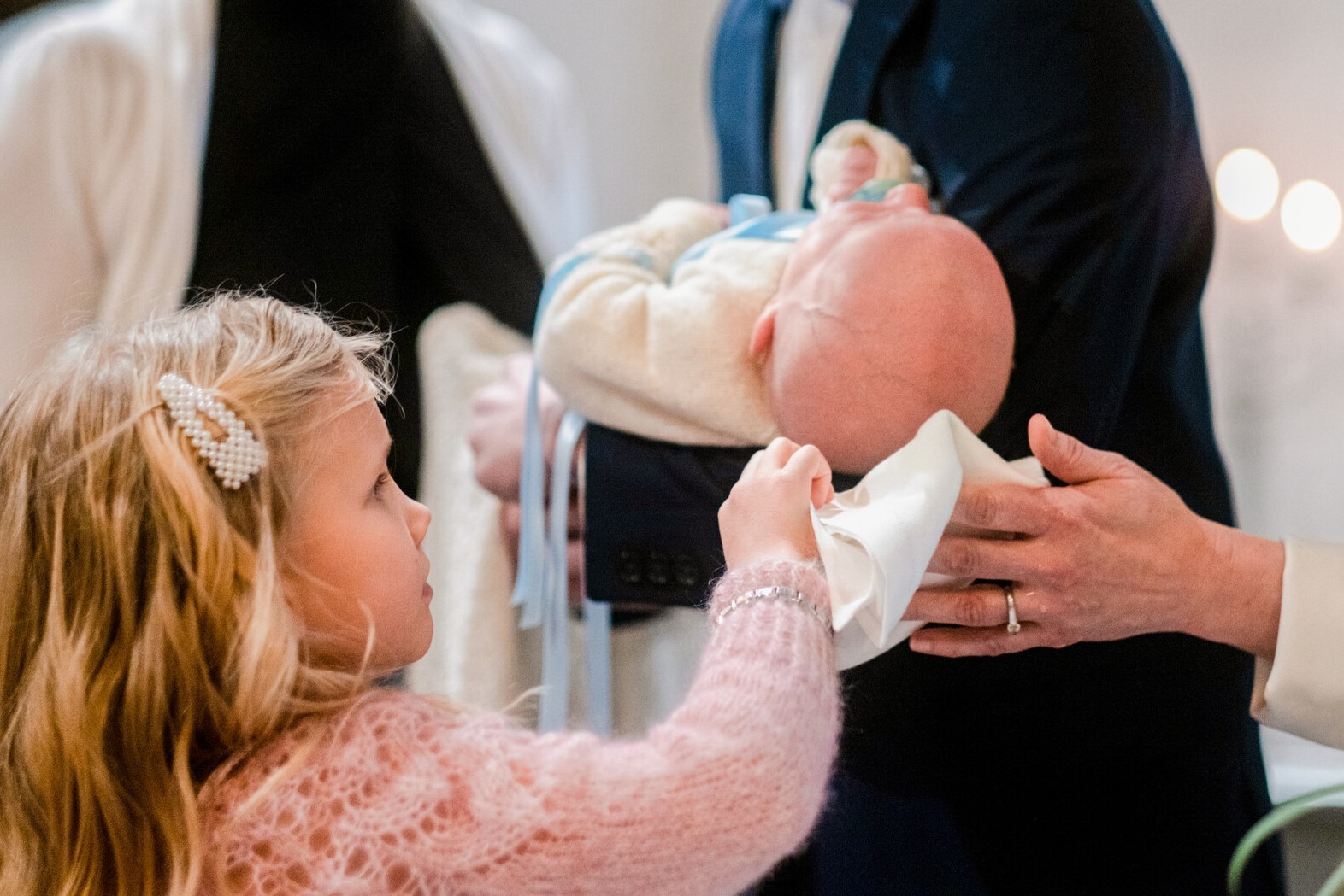 Pieni lapsi kuivaa vauvan päätä liinalla, jota pitää myös papin käsi ja taustalla vauvaa sylissä pitävä aik...