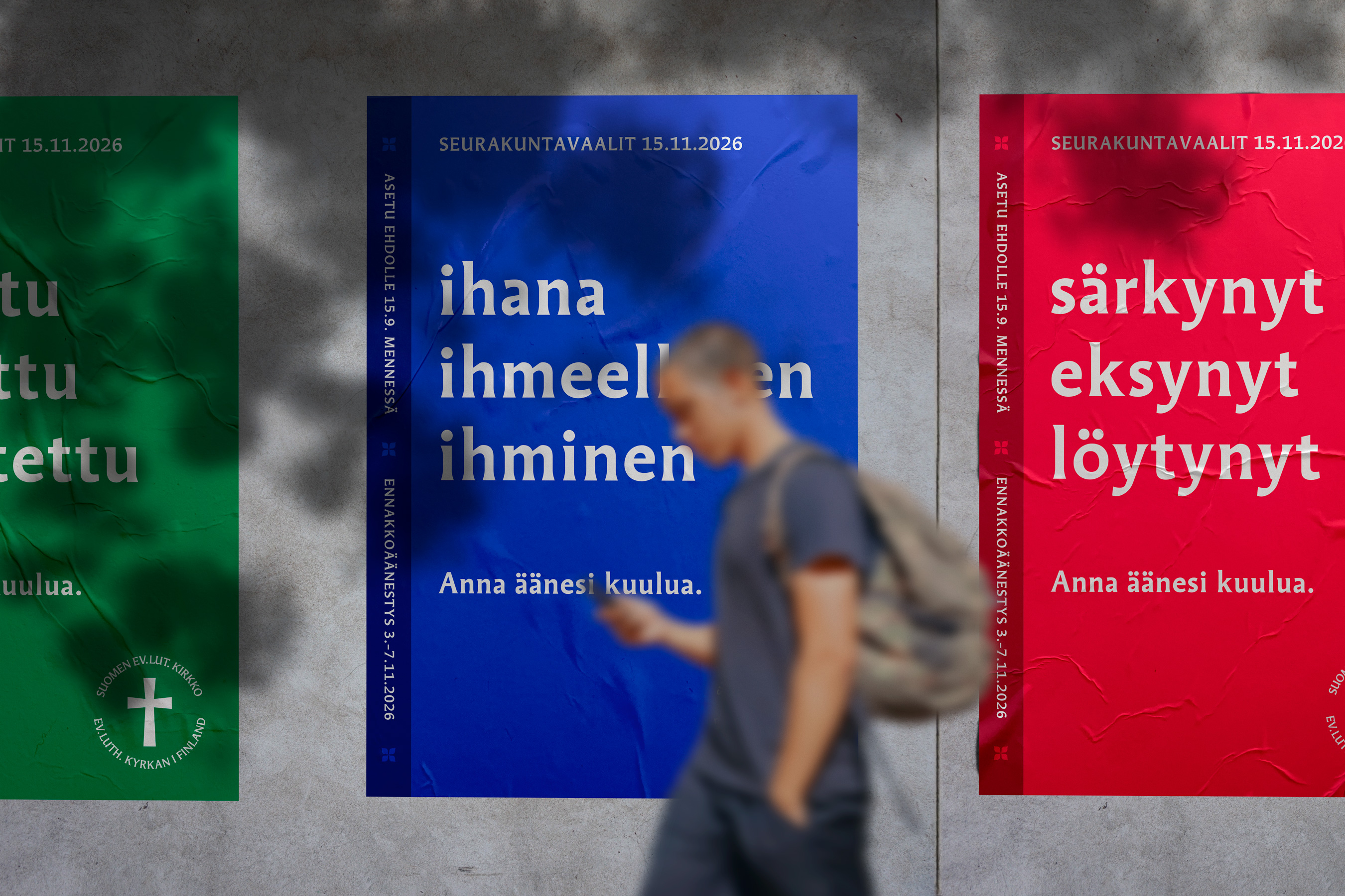 Kolme julistetta seinällä, joissa on eri värejä ja tekstejä. Henkilö kävelee ohi, taustalla näkyy punainen,...