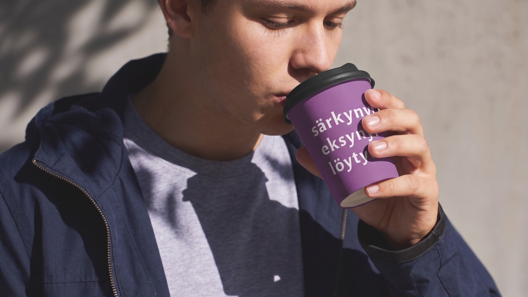Mies juo kahvia violetista kupista, jossa on tekstiä. Hänellä on sininen takki ja harmaa paita.