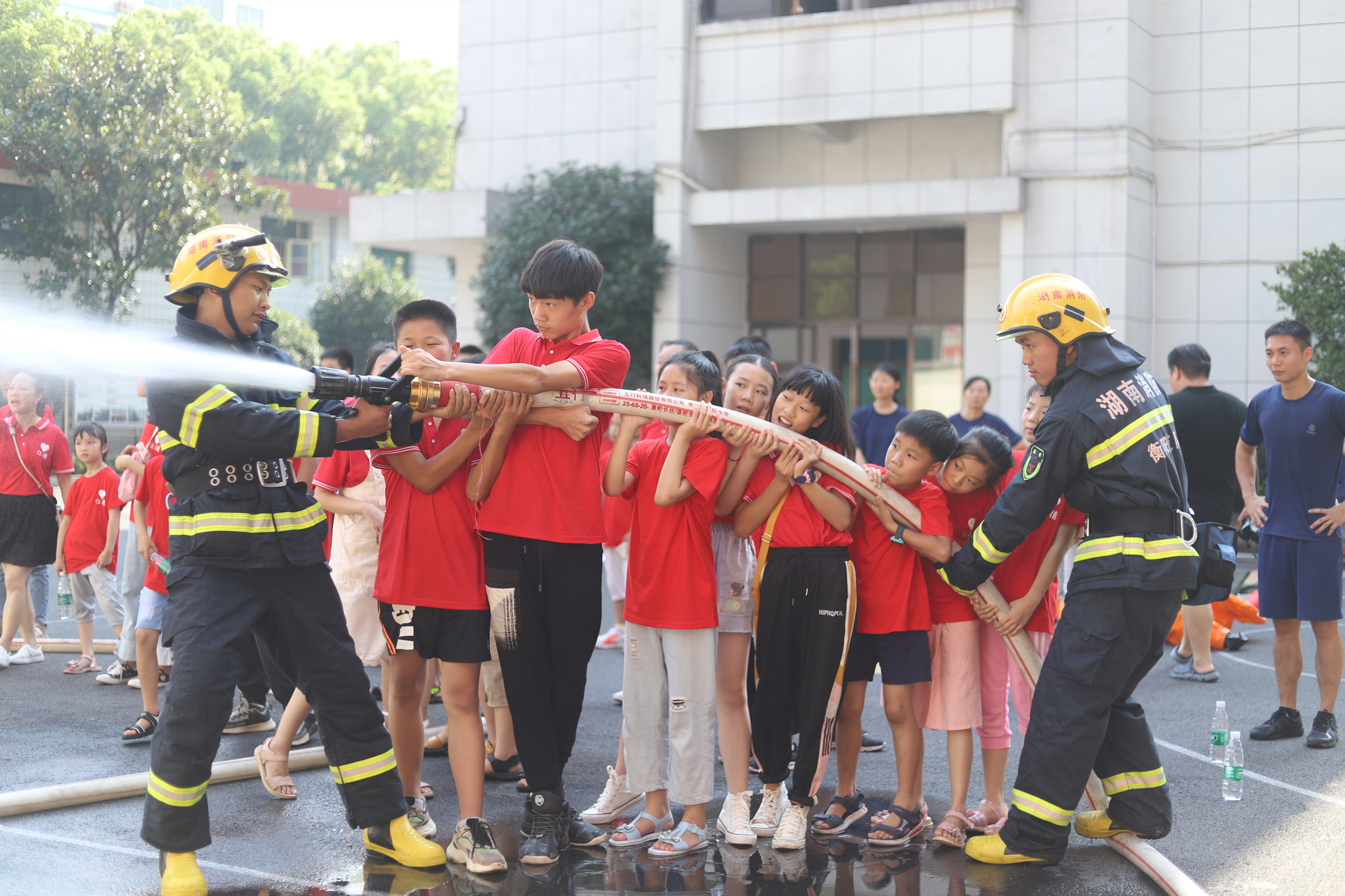 Kiinalaisia lapsia ja palomiehiä ruiskuttamassa vettä paloletkusta.