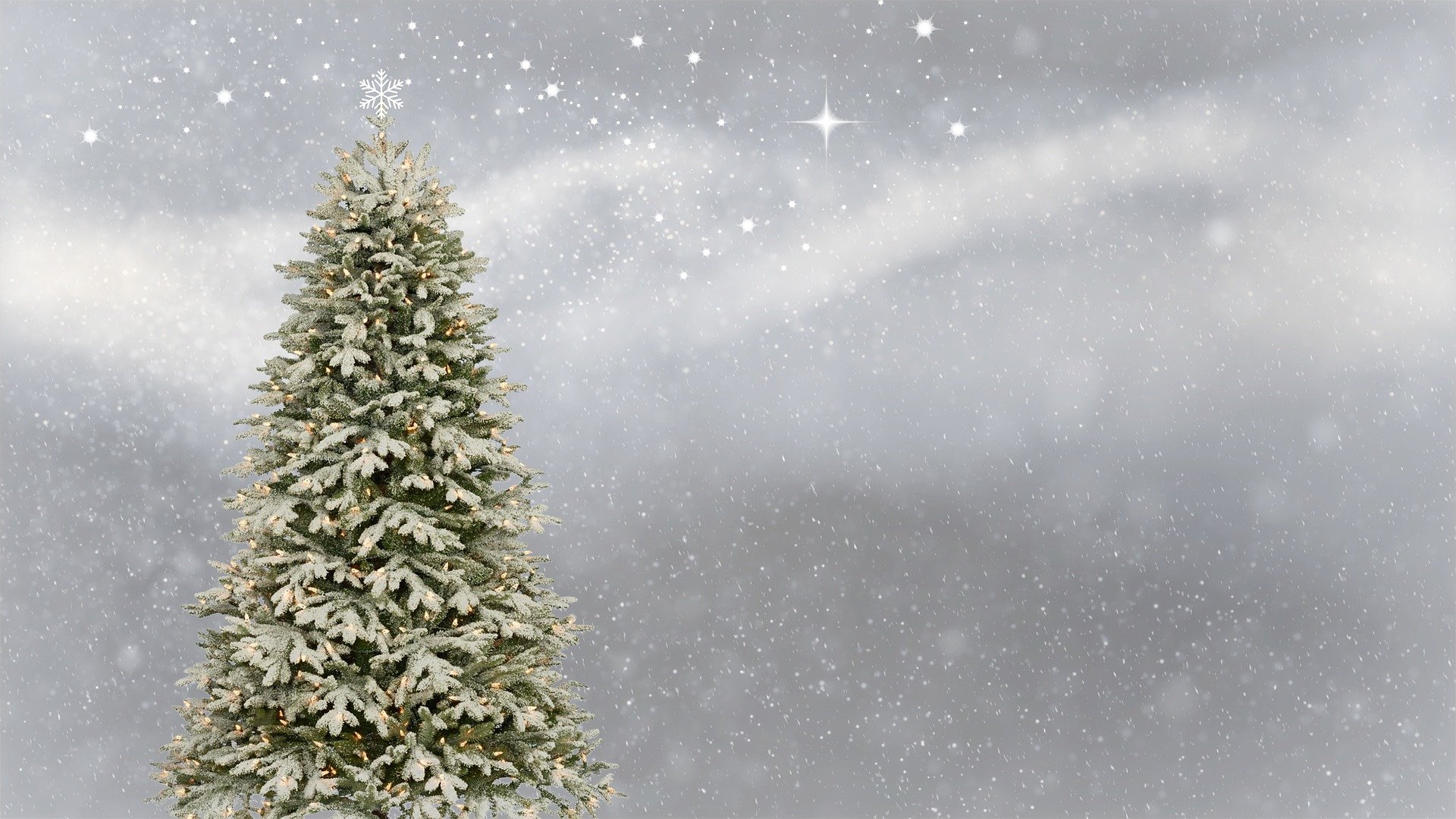 Hennosti lumipeitteinen kuusi ja siinä kynttilän valoja ja latvassa valkoinen joulutähti ja siniharmaalla taustalla kevyttä lumisadetta.