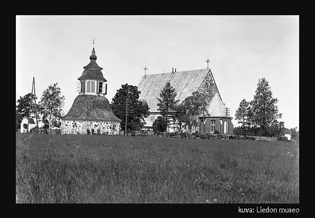Mustavalkoisessa vanhassa kuvassa Liedon kirkko kiviaitoineen kuvattuna Kirkkotien suunnalta pellon takaa ja aidan edessä on kirkkoväkeä  hevosineen.