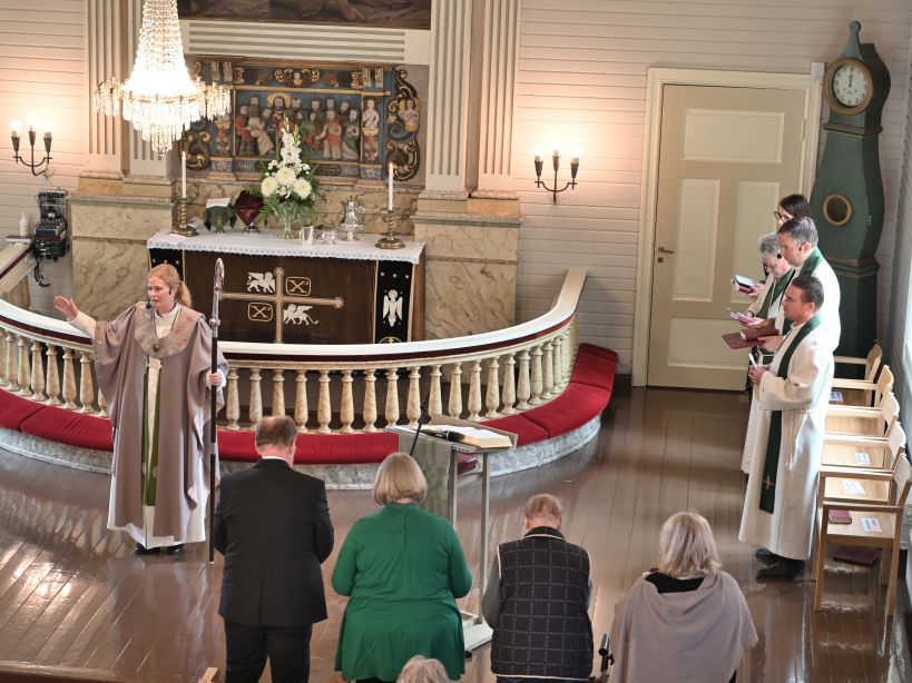 Alttarialueella piispa siunaa ja avustajia istuu sivulla ja kirkon etupenkissä.