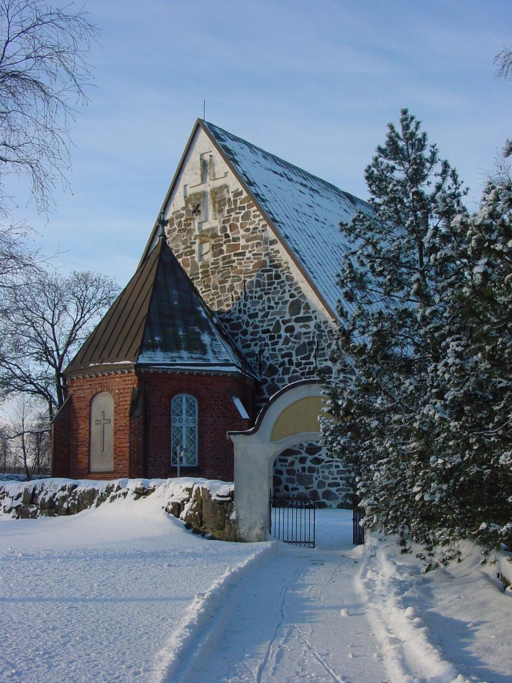 Papin portti avoimena lumisessa talvimaisemassa sekä edessä lumesta luotu kulkuväylä ja takana Liedon kirkko.