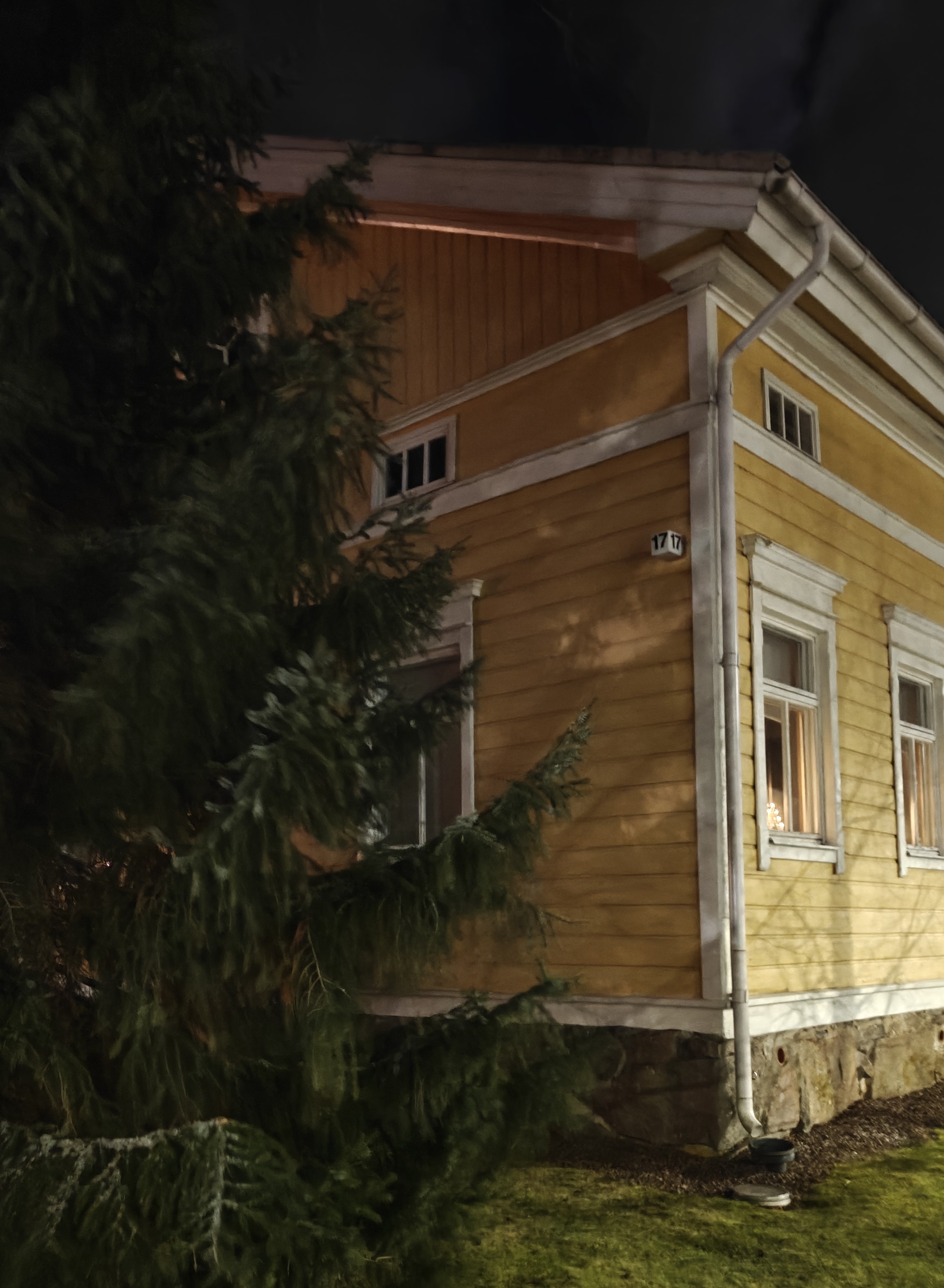 Kuusipuu ja keltaisen puutalon kulmaa, ikkunoissa kynttelikön valoa, maassa ruohoa ja tummaa taivasta.