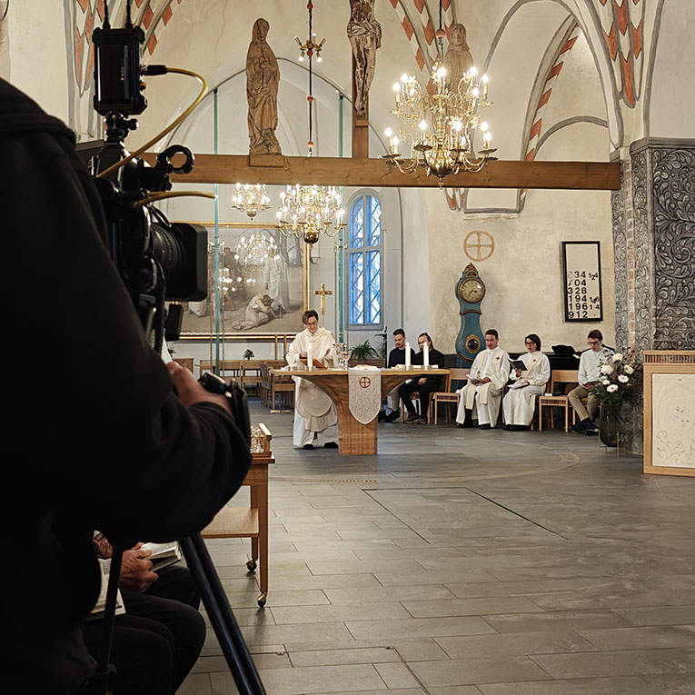 Tv-kamera kuvaa Liedon kirkkosalia jossa pappi alttarilla ja alttarin sivussa ja takana tuoleilla istumassa...