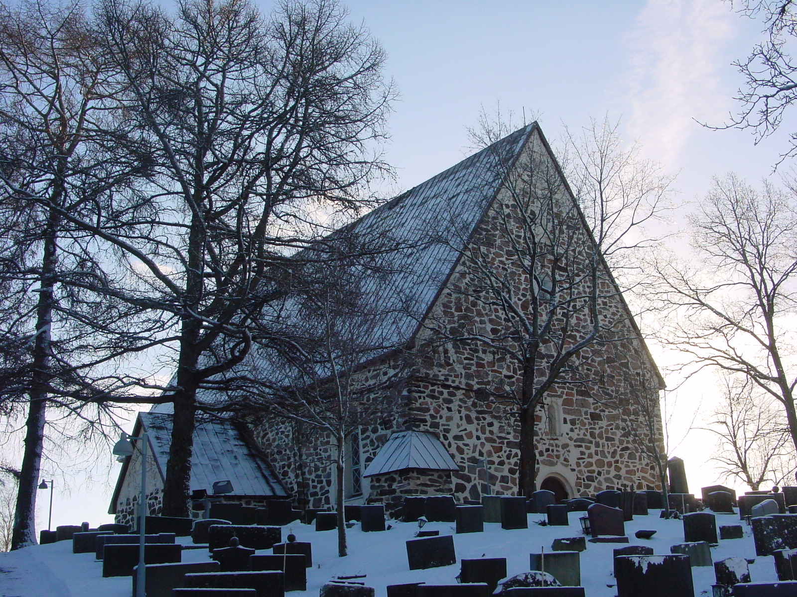 Luonnonkivistä muurattu kirkko, jossa on korkea harjakatto. Edustalla on paljon hautoja. Ympärillä on lunta.