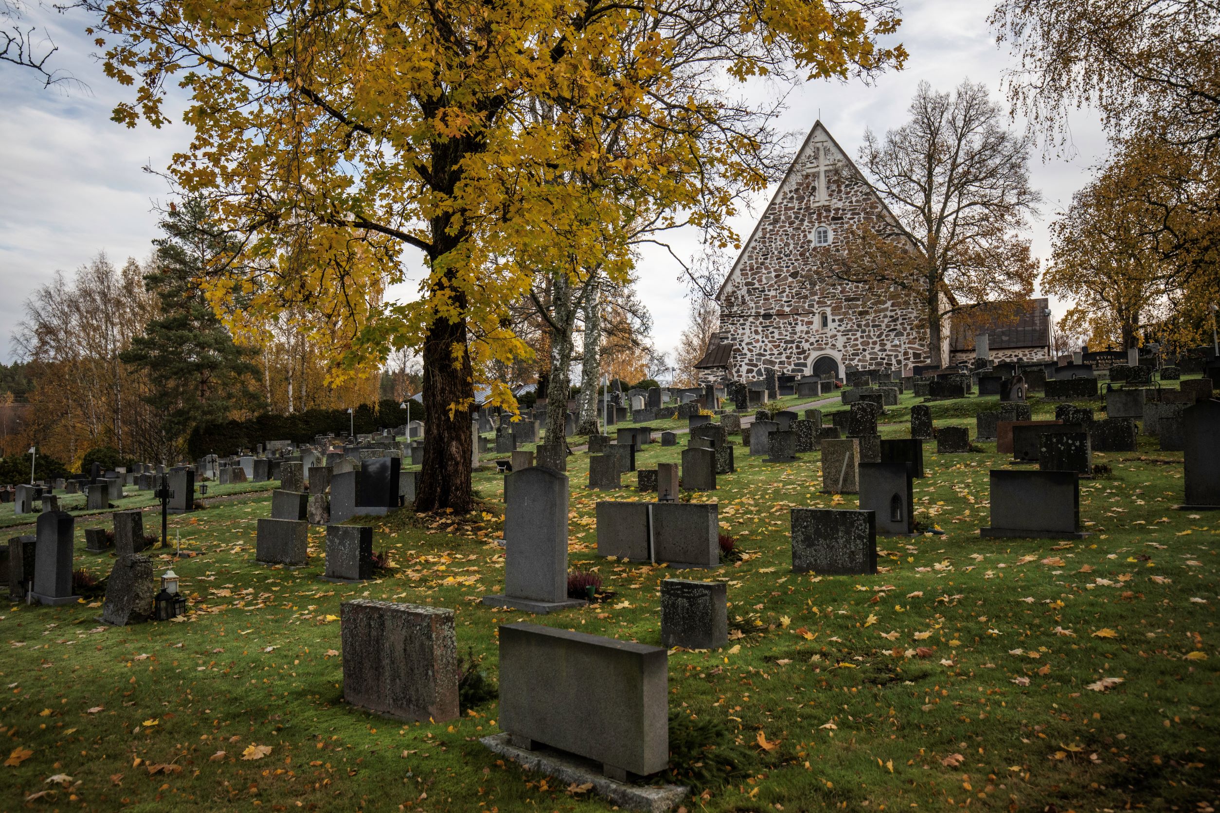 Useita hautoja rinteessä, joiden keskellä on ruskainen lehtipuu. Kuvan takaosassa hautausmaan kummun päällä näkyvät kivinen kirkko ja sen asehuone. Maassa on paljon lehtiä.