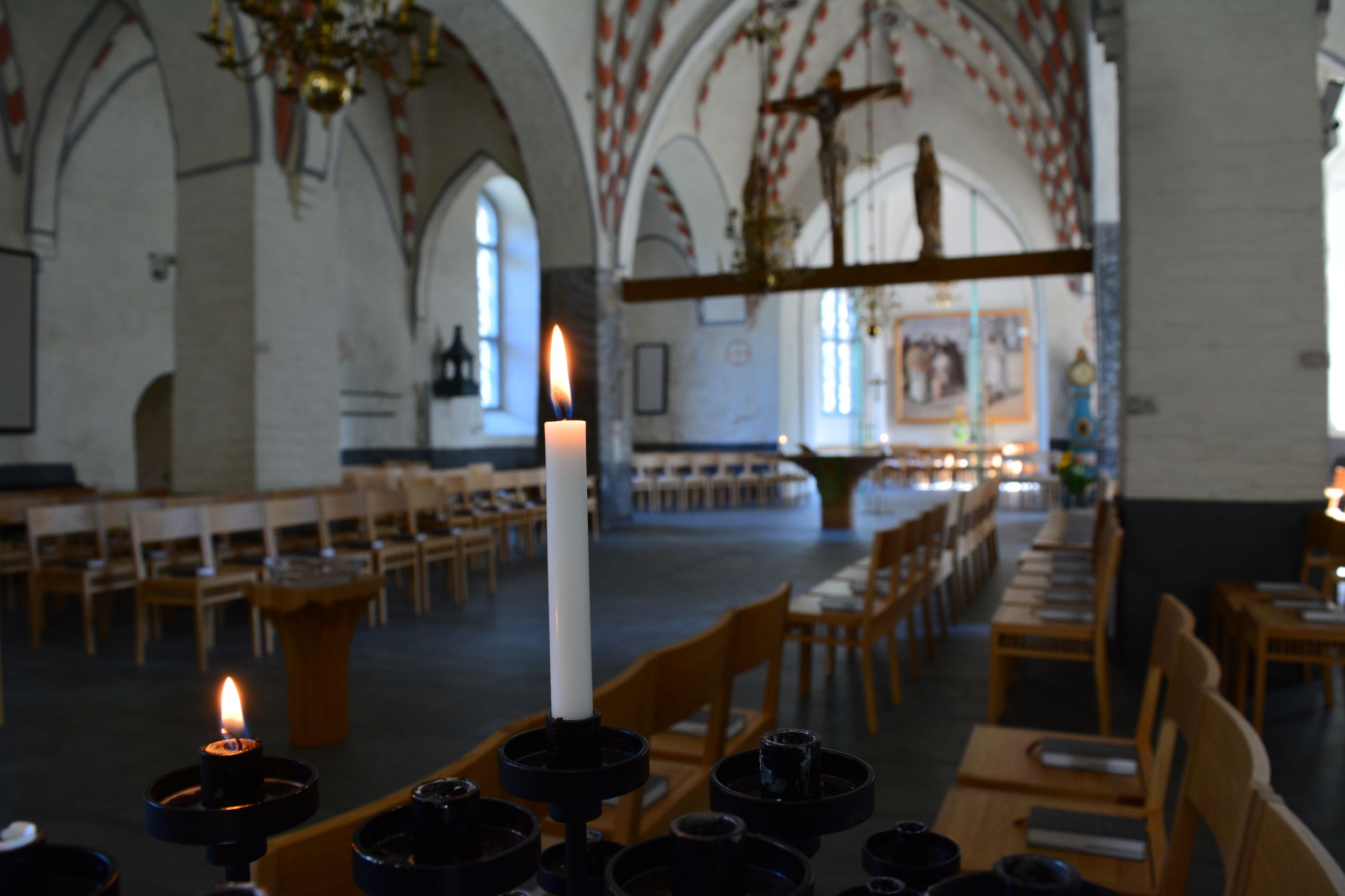 Mustassa ja rautaisessa kynttelikössä palaa kaksi pientä kynttilää hämärässä kirkkosalissa. Taustalla näkyvä muu sali on sumea, ja kuva on kohdistettu kynttilöihin.