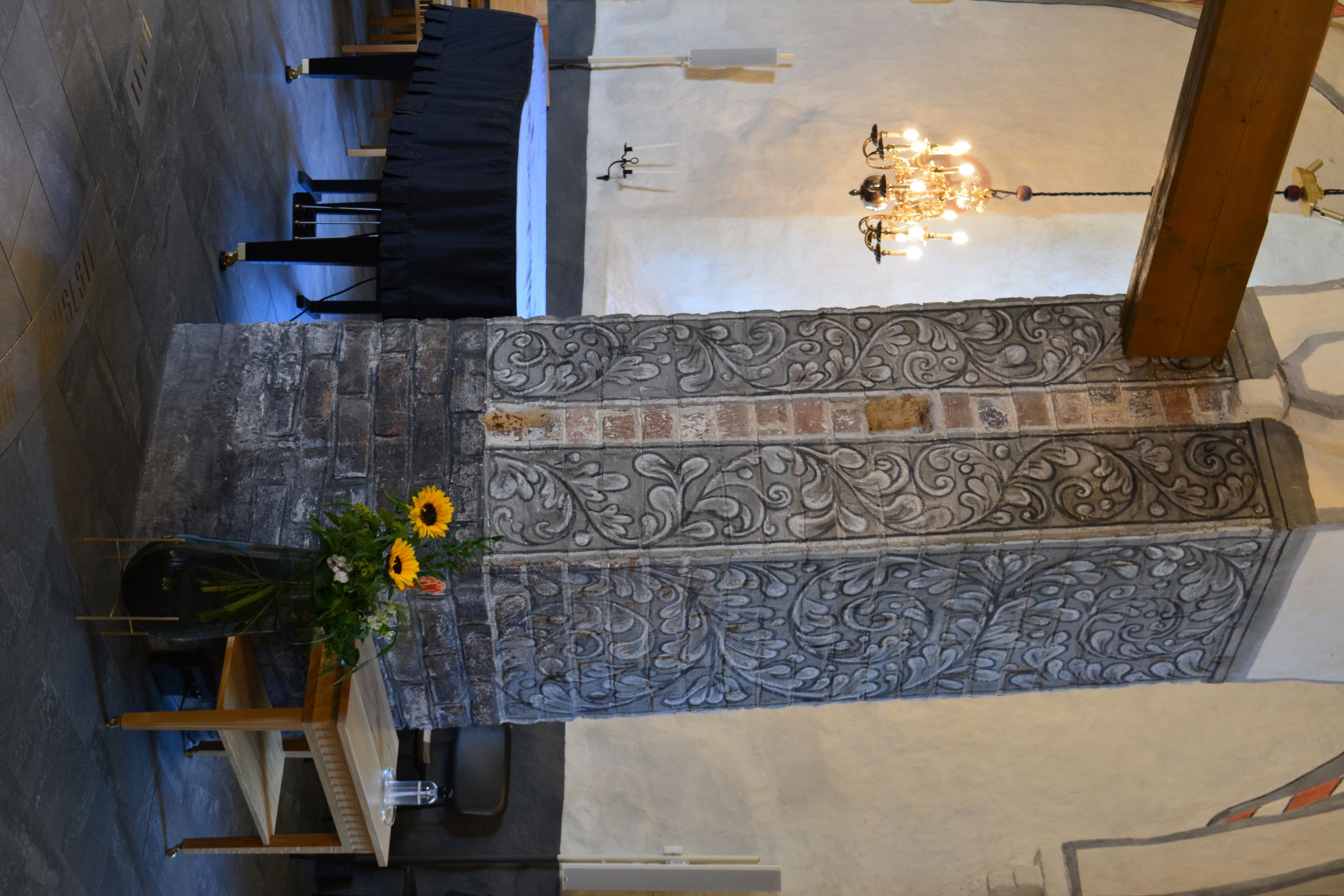 Tiilistä rakennettu paksu pilari, jonka pintaan on maalattu harmaan eri sävyillä köynnöskuviota.