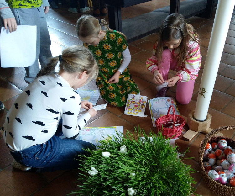 Lasten pääsiäinen Lapsia lattialla askartelemassa. Etualalla runsaasti vihreää ruohoa ja sivulla suklaamunia korissa.