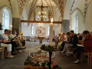 Etualalla palava kynttilä ja kastemalja ja sen takana väkeä Liedon kirkkosalissa istumassa ja veisaamassa. 