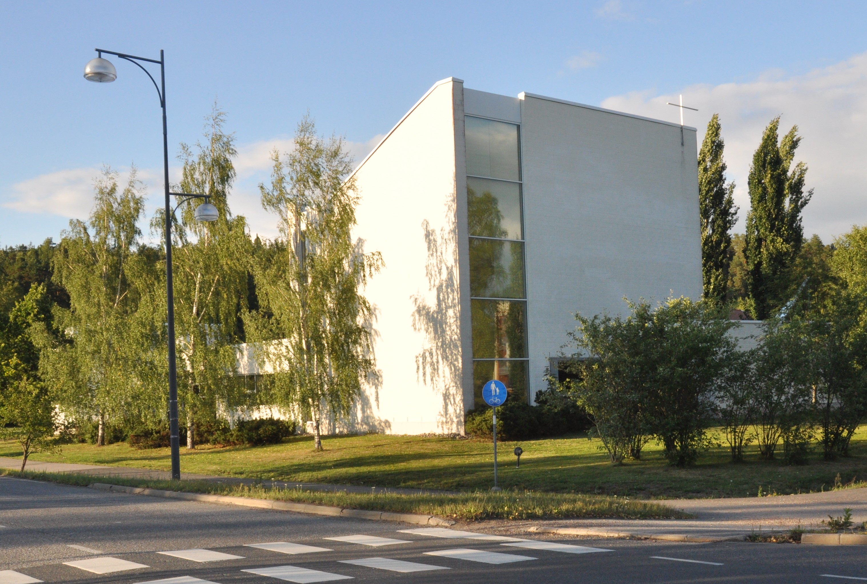 Liedon seurakuntatalo: valkoinen 90-luvun tiilikivitalo päätyristeineen ja korkeine päätyikkunoineen kadulta päin katsottuna auringonvalossa. 