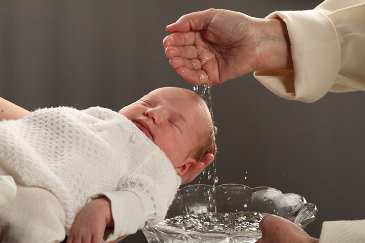Käsi valelee vettä kastevettä sylissä olevan vauvan päähän.