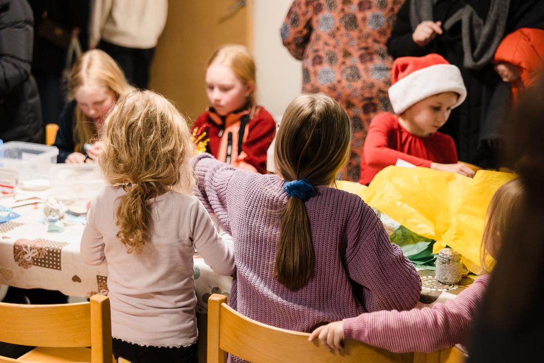 Lapset askartelevat joulukoristeita pöydän ääressä, taustalla näkyy muita ihmisiä juhlatunnelmassa.