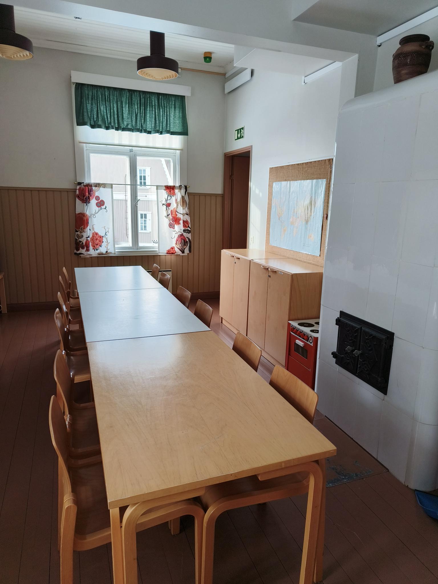 Pieni huone, pöytiä, tuoleja, kaakeliuuni, hyllykkö, lasten hella.